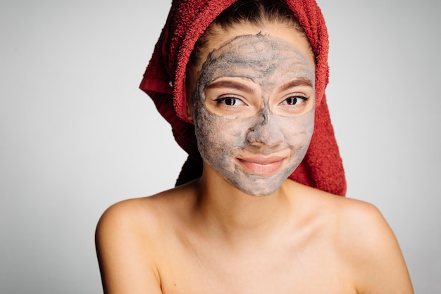 Charming young girl with a red towel on her head wants to be beautiful on her face a useful clay mask