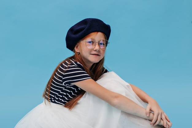 Charming young lady with long hair and freckles in striped white and blue clothes looking into camera on isolated background