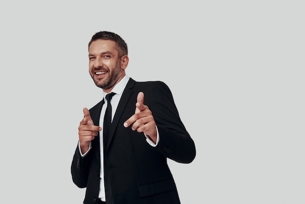 Charming young man in full suit smiling and pointing you while standing against grey background