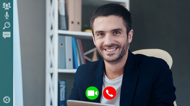 Charming young man in smart casual wear looking at camera and smiling while having video call