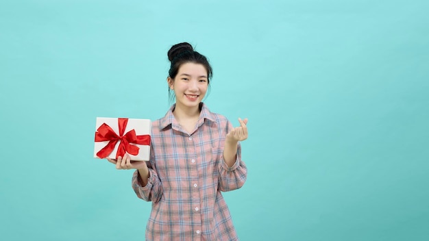 Charming young woman Asian with dark long hair feeling happy smiling to the camera and makes Korean love sign shapes heart, with fingers and holding gift box isolated over blue background.