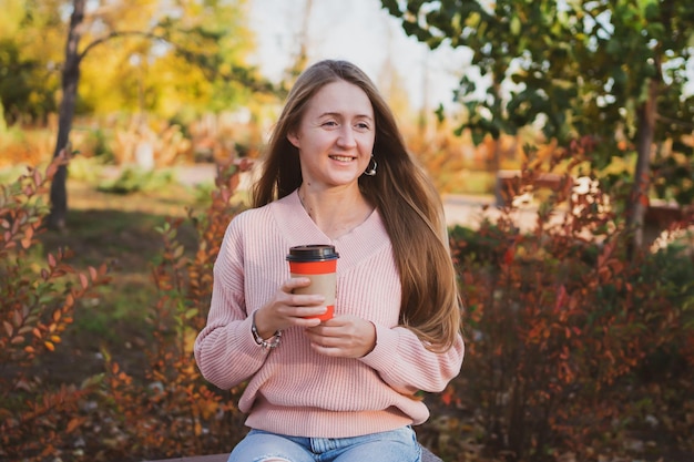 Charming young woman drinks coffee sitting on bench in beautiful autumn park