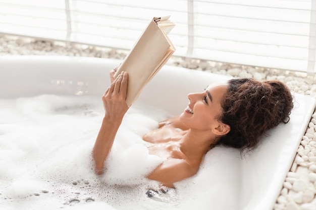 Charming young woman lying in bubble bath reading book in relaxing atmosphere at home free space