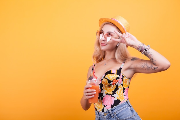 Charming young woman making peace signs while having a fresh orange juice with sunglasses over yellow bacground in studio