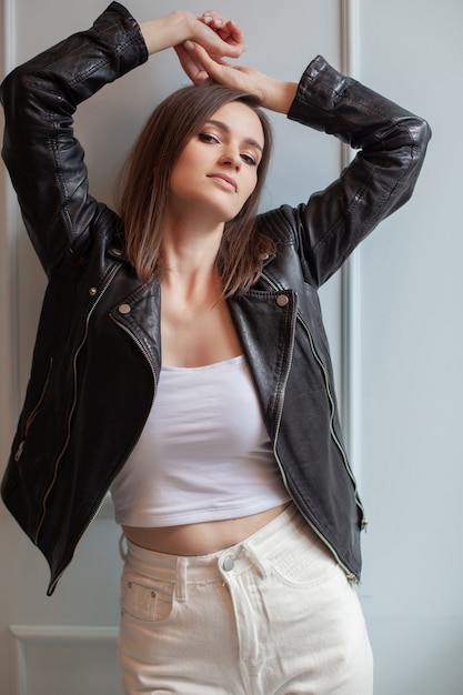 Charming young woman posing in black leather jacket and white jeans on a white background. Beauty, fashion.