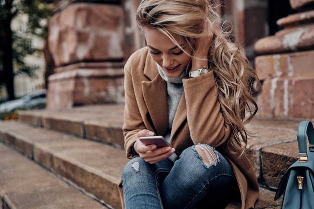 Chatting with friends. Beautiful young woman using her smart phone and smiling while sitting on stairs outdoors