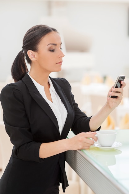 Checking business messages. Confident young woman in formalwear drinking coffee and looking at her mobile phone while leaning at bar counter