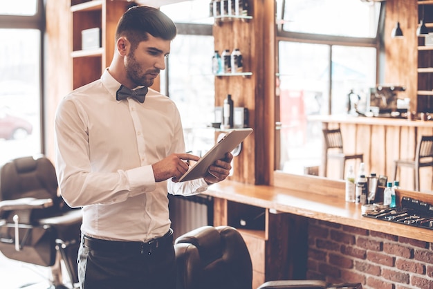 Checking clients list. Young handsome man using his digital tablet while standing at barbershop