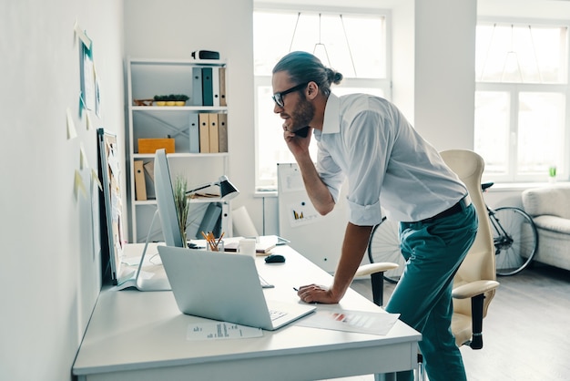 Checking every detail. Good looking young man in shirt talking on the smart phone and smiling while working in the office