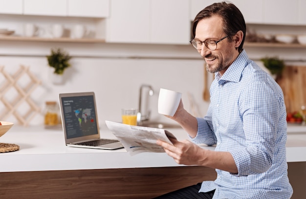 Checking yellow press. Gifted creative smiling self employed businessman sitting at home and drinking juice while using gadget and reading newspaper