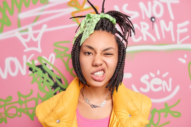 Cheeky hipster girl with combed dreadlocks clenches teeth smirks face looks self confident at camera spends free time at street poses over graffiti wall wall