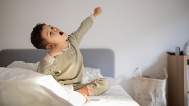 Cheerful active caucasian little child wake up sits on bed stretching body yawns in bedroom interior panorama