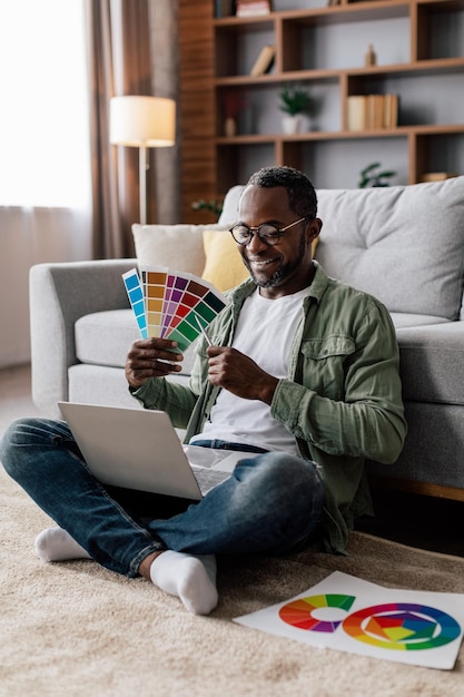 Cheerful adult black guy designer in glasses and casual show color palettes in laptop on floor in living room