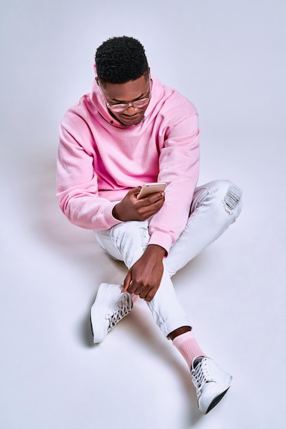 Cheerful african american guy using phone sitting floor at studio on grey background Vertical photo