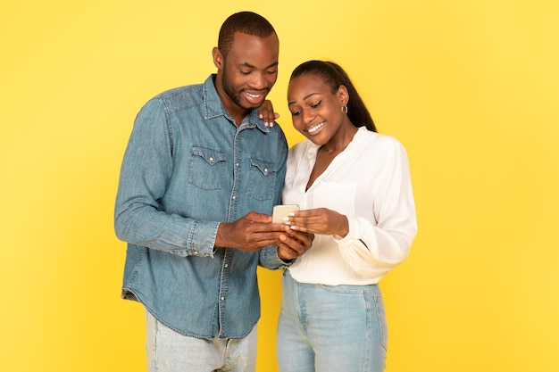 Cheerful African American Spouses Using Smartphone Together Over Yellow Background