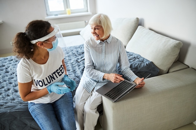 Cheerful aged female with a laptop on a bedside listening to a care worker explanation about a thing on display