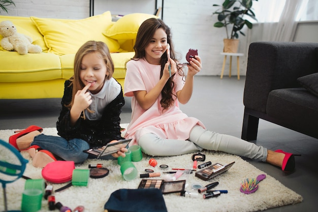 Cheerful ans exited girls sitting on floor in room. They doing makeup. Teenagers look in mirror and use cosmetics.