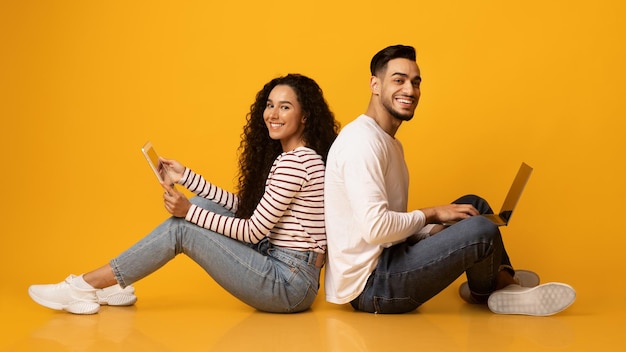 Cheerful arab couple sitting back to back with laptop and digital tablet