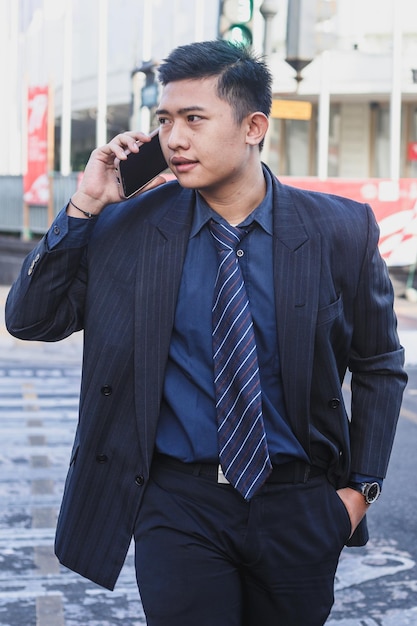 Cheerful Asian businessman making a phone call and smiling while walking outside in the city