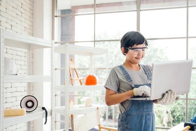 A cheerful Asian carpentry boy wearing safety goggles and work gloves working on a laptop in the home workshop studio Construction work and repairs Carpentry and family concept