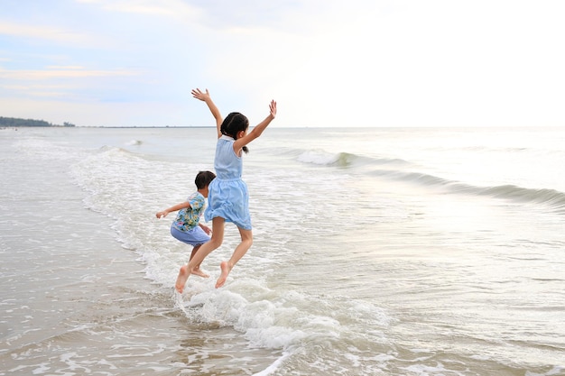 Cheerful Asian child girl and little boy having fun jumping on wave at tropical sand beach at sunset Happy family enjoy in summer holiday