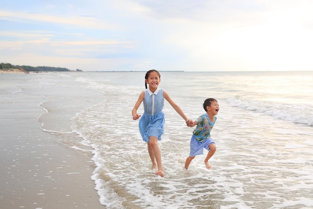 Cheerful Asian child girl and little boy having fun run together and hand in hands on tropical sand beach at sunset Happy family enjoy in summer holiday