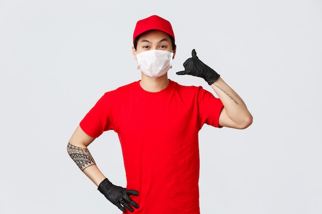 Cheerful asian delivery guy in medical mask and protective gloves show phone sign, asking clients give call anytime when delivery needed.