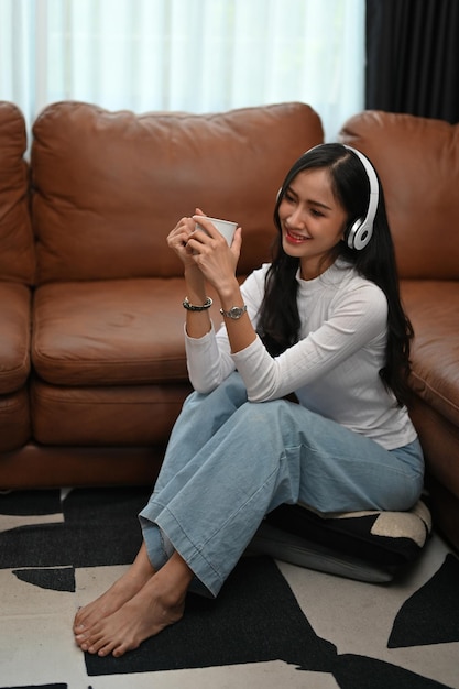 Cheerful Asian female listening to music sipping coffee and sitting on then living room floor