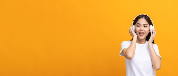 Cheerful asian female teenager listen to the music with white headphone dancing on isolated yellow background. Beautiful young woman in hand touch a wireless headphone having fun with music.