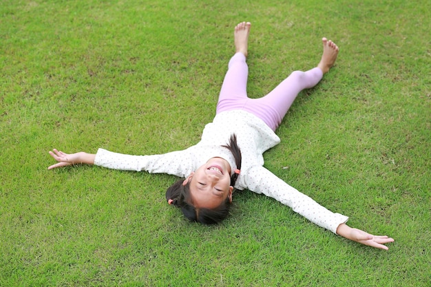 Cheerful Asian girl child lying down on green lawn Happy kid lies on grass and looking at camera