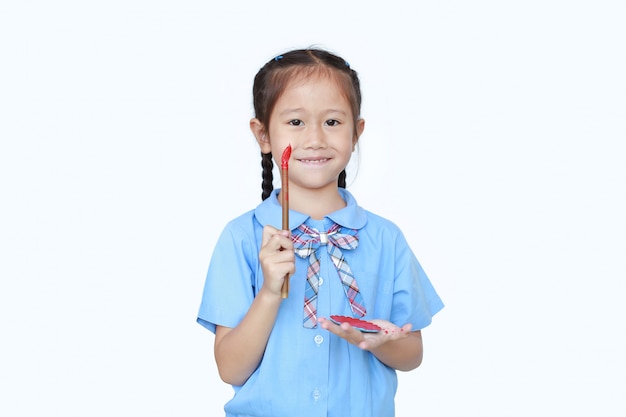 Cheerful Asian little girl in school uniform holding Red dipped paintbrush and palette