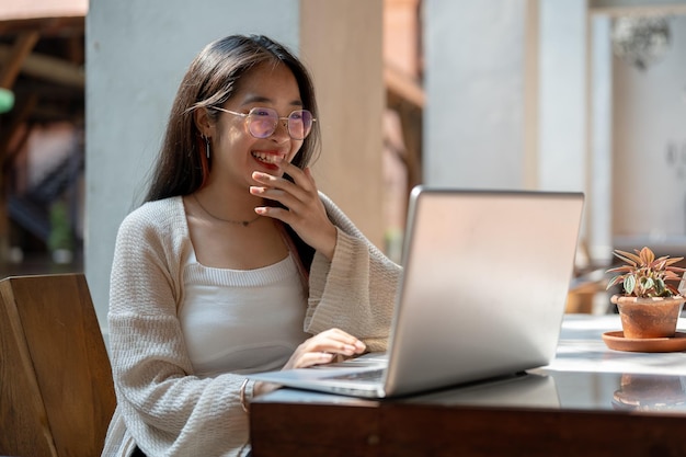 A cheerful Asian woman is laughing while enjoying talking on a video call on her laptop at a cafe