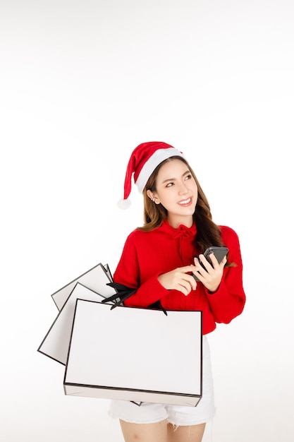 Cheerful attractive Asian woman wearing a red dress with a Santa hat is holding a shopping bags on white isolated background. Beautiful lady with long black hair is using cell phone shopping online.