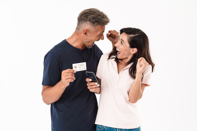 Cheerful attractive couple wearing casual outfit standing isolated over white wall, shopping online with mobile phone and credit card