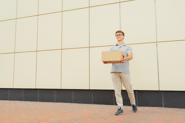 Cheerful attractive courier in eyeglasses, looking while holding a delivery box and clipboard, standing against the wall, outdoors.