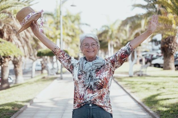 Cheerful attractive elderly woman standing in public park with outstretched arms holding her hat looking at camera