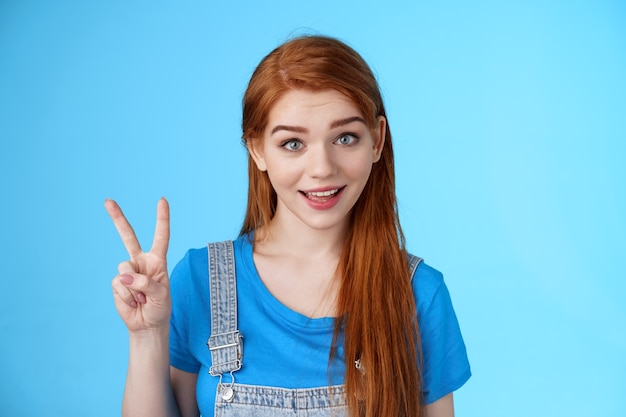 Cheerful attractive redhead caucasian girl smiling joyfully talking, order two peaces, show second number, peace victory sign, stand carefree, upbeat emotions, stand blue background