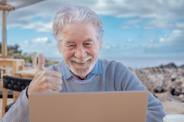 Cheerful attractive senior man video calling by laptop computer by the beach Elderly bearded male sitting at a wooden table close to the sea