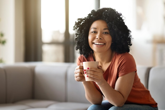 Cheerful attractive young african american woman chilling alone at home drinking coffee