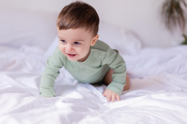 Cheerful baby in a green cotton bodysuit is lying on his stomach on a white bed