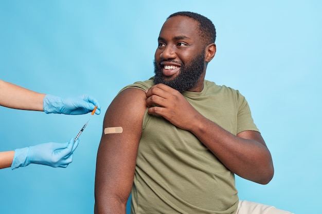 Cheerful bearded adult Afro American man got covid 19 vaccine looks with happy expression at nurse shows shoulder with adhesive tap poses against blue wall