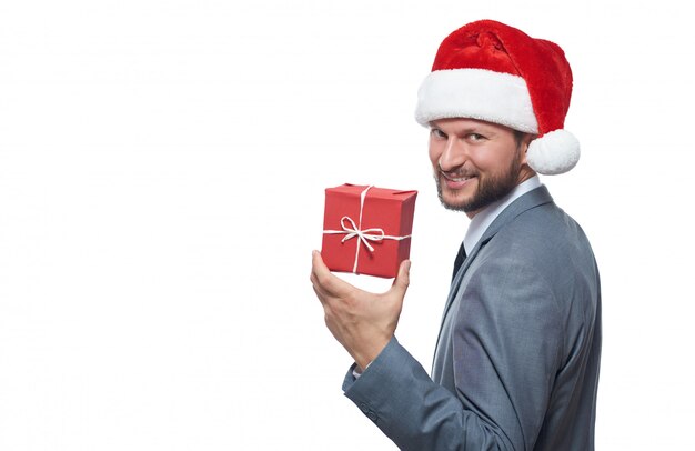 Cheerful bearded businessman in Christmas hat smiling over shoulder keeping small Christmas gift.