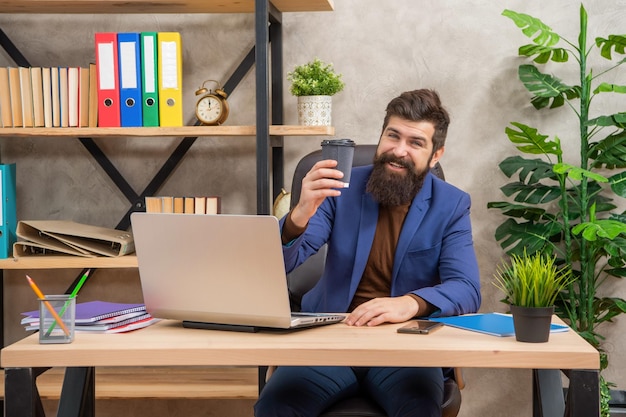 Cheerful bearded businessman work on laptop with coffee in the office modern life