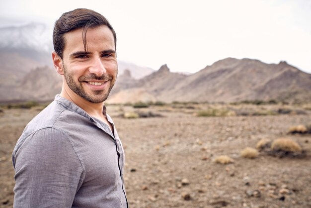 Cheerful bearded male traveler in casual clothes standing on dry grassy stony arid terrain and smiling looking at camera