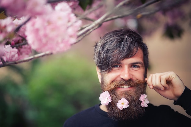 Cheerful bearded man with long mustache and sakura flowers in beard smiling near trees