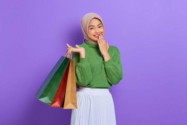 Cheerful beautiful Asian woman in green sweater holding shopping bags with hand on mouth isolated over purple background