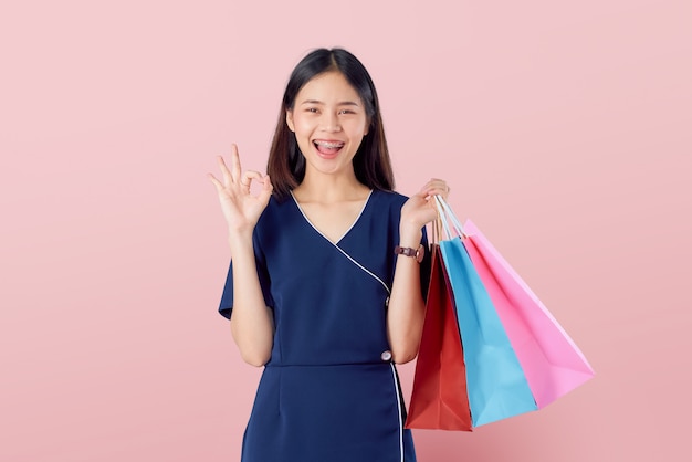 Cheerful beautiful Asian woman holding multi coloured shopping bags and shows ok sign on light pink wall