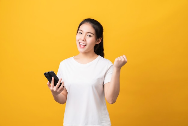 Cheerful beautiful Asian woman holding smartphone on light yellow background.