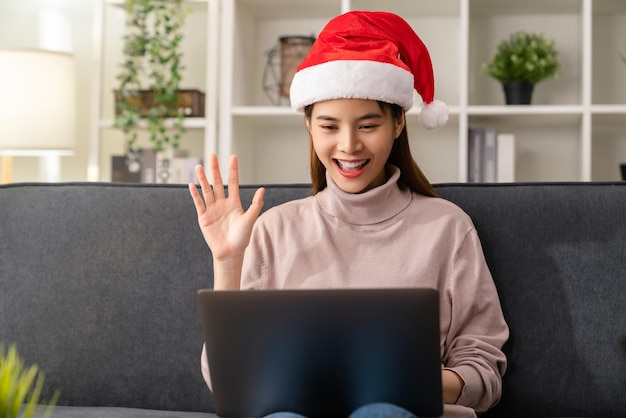 Cheerful beautiful Asian woman wearing red christmas hat and video conference online talking with friends to laptop on the sofa at house.