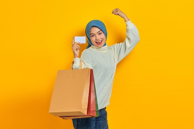 Cheerful beautiful Asian woman in white sweater holding shopping bag and credit card celebrate luck isolated over yellow background